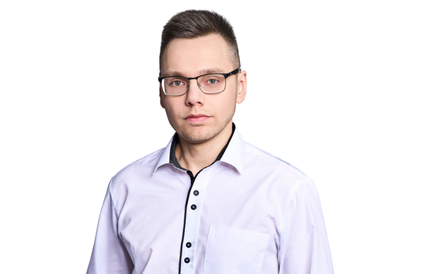The photo of the article's author - our front-end developer. The picture shows a professional young man with short hair and rectangular glasses looks directly at the camera with a serious expression. He is wearing a smart lavender shirt with black buttons, suggesting a business-casual attire suitable for a modern office environment. The background is plain white, focusing all attention on him.