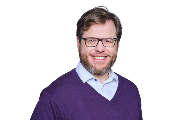 Portrait of Bernhard Huber, Primotly's Founder, wearing glasses, a purple sweater over a light blue shirt, and showcasing a warm, engaging smile. His professional yet approachable demeanor is captured against a plain white background, ideal for accompanying his authored articles and tech discussions